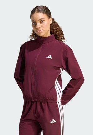 Femme aux cheveux bouclés portant un survêtement Adidas bordeaux à rayures blanches, posant les mains sur les hanches devant un fond uni.