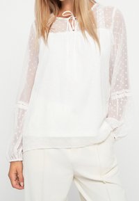 Blusa bianca in tessuto semitrasparente a pois, maniche lunghe a sbuffo, collo a chiave con un laccio e dettagli in pizzo ai polsini.