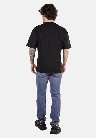 T-shirt nera in cotone con maniche corte, indossata con jeans azzurri. La persona ha i capelli ricci e tatuaggi, voltata di spalle rispetto alla fotocamera.