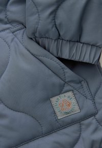 Veste matelassée en tissu bleu marine avec des poignets élastiques. Comprend une étiquette carrée avec un logo brun et du texte, ainsi que des motifs matelassés arrondis.
