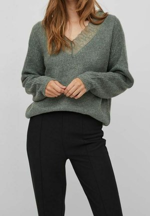 Kvinde iført en løs grå strikket sweater over en grøn top med blondekant og tætsiddende sorte bukser mod en ensfarvet baggrund.