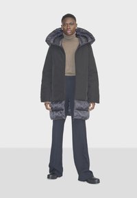 Dlhý čierny zateplený kabát s prešívanou kapucňou a rukávmi, so zipsovým uzáverom a dvoma bočnými vreckami, doplnený o tmavé nohavice a topánky.