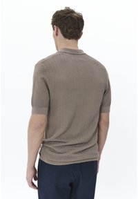 Polo de manga corta marrón con un patrón de zigzag texturizado, cuello y puños acanalados. Lleva puesto un pantalón azul marino, de espaldas a la cámara.
