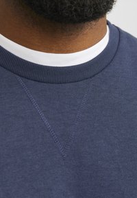 Navy blauwe sweatshirt met een geribbelde ronde hals. Voorzien van een V-stiksel detail aan de voorkant en een witte binnenkraaglaag. Gladde textuur.