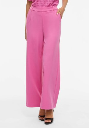 Kobieta ubrana w różowe spodnie z wysokim stanem i szerokimi nogawkami z bocznymi kieszeniami oraz czarne sandały na obcasie z odkrytymi palcami, stojąca na białym tle.