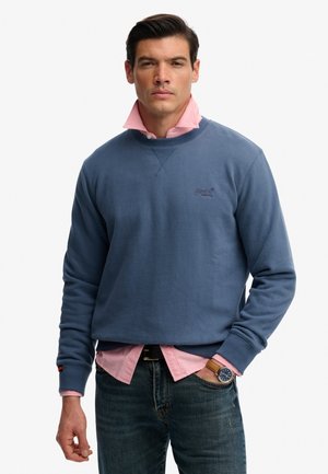 Man draagt een blauwe crewneck trui over een roze overhemd met kraag, blauwe spijkerbroek en een polshorloge, staand met één hand in de zak.