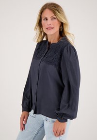 Navyblauwe blouse met kantdetails op de schouders, knoopsluiting aan de voorkant en uitlopende lange mouwen, gemaakt van een gladde stof.
