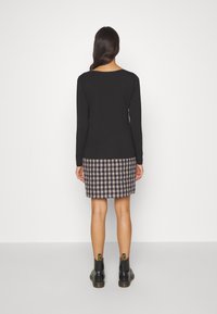 Kvinna som står med ryggen mot kameran, iklädd en svart långärmad topp, rutmönstrad kjol och svarta ankelboots framför en vit bakgrund.