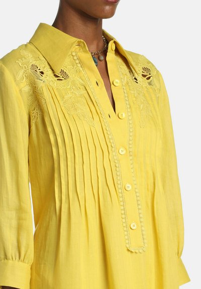 Žltá blúzka s dlhými rukávmi, s kvetinovou čipkovou výšivkou na ramenách, vertikálnymi záhybmi vpredu, zapínaním na gombíky a špicatým golierom.