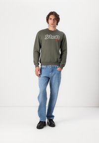 Felpa verde oliva con logo "Schott NYC" bianco e rosso, abbinata a jeans azzurri. Il modello indossa scarpe nere, in piedi su uno sfondo bianco.