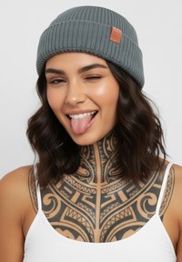 Gray ribbed knit beanie with a leather patch; model showing a tattooed neck and sticking out her tongue, wearing a white tank top.