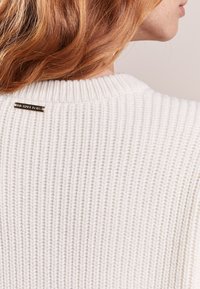 Biały sweter z dzianiny o ryflowanej fakturze, z zapięciem z tyłu z metalowym akcentem oraz okrągłym dekoltem.