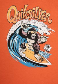 Oranžové tričko s grafikou gorily surfařící, nosící sluneční brýle, se stylizovanými vlnami a textem "Quiksilver" nahoře.