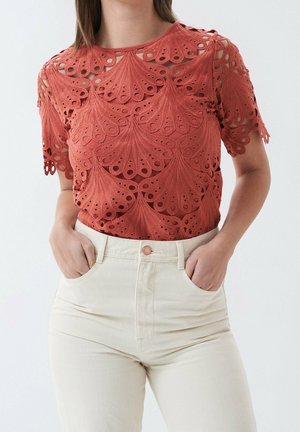 Koronkowa bluzka w kolorze rdzawego z krótkimi rękawami i falowanymi brzegami, noszona z wysokim stanem białawych spodni, ręce włożone do kieszeni.