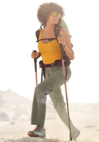 Wandererausrüstung: gelbes Tanktop, grüne Hose, robuste Stiefel, schwarzer Rucksack, Trekkingstöcke. Helle Sonnenbrille, natürliche Umgebung.