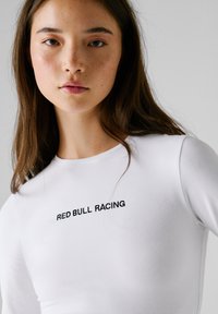 Młoda kobieta z długimi brązowymi włosami, ubrana w białą koszulę z napisem "RED BULL RACING" na piersi, na jednolitym jasnym tle.