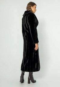Dlouhý černý kabát z umělé kožešiny s širokým límcem, má rovný střih a hladkou strukturu. Nosit se nad hnědými koženými botami na podpatku.