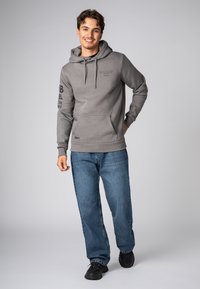 Grijze hoodie met een buidelzak, capuchon met drawstring en opdruk op de mouwen. Gecombineerd met blauwe spijkerbroek en zwarte sneakers.