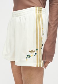 Λευκό σορτς με χρυσές ρίγες και κεντήματα με λουλούδια, συνδυασμένο με μακρυμάνικη καφέ μπλούζα στο κάτω μέρος σώματος ενός όρθιου ατόμου.