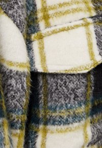 Ullpläd i grå, vit och gul färg med en mjuk, fluffig textur. Designen inkluderar ett kontrasterande rutmönster och sydda kanter.