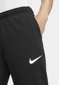 Zwarte sportbroek met een elastische tailleband, zijzakken en een wit Nike-logo op de linkerdij, gemaakt van een zachte, gestructureerde stof.