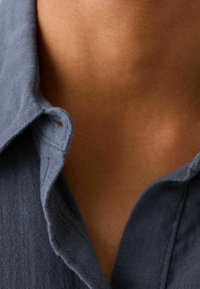 Šedý texturovaný límec košile s knoflíkovou dírkou, zobrazený na odhalené kůži. Měkká tkanina a volný střih. Viditelný výstřih.