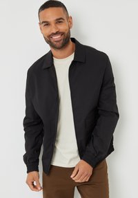 Giubbotto bomber nero con chiusura a zip, colletto piatto, tasche laterali e polsini a costine. Indossato sopra una maglietta beige a girocollo.
