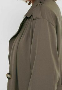 Oliwkowy trencz o luźnym kroju, z zapięciem na guziki, akcentami na ramionach oraz gładkim, teksturowanym materiałem.