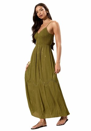 Mujer vistiendo un vestido maxi sin mangas de color verde oliva con tirantes finos, cuerpo fruncido y un sutil detalle de brillo vertical, de pie con una mano sujetando la falda.