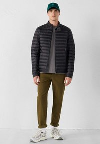 Giacca nera imbottita con colletto alto, camicia grigia sotto, pantaloni verde oliva e sneaker grigi. Il cappellino è color teal con un dettaglio del logo.