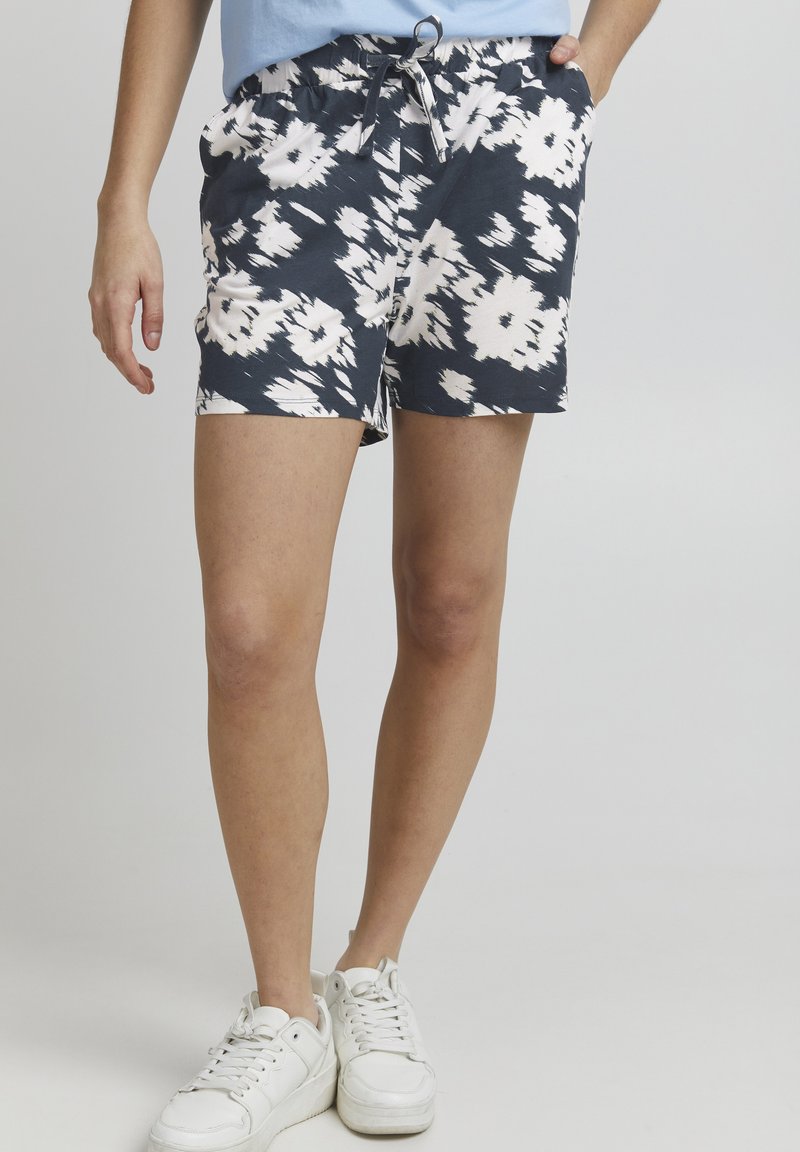 Shorts à motif floral en bleu marine et blanc, fabriqués en tissu léger. Dotés d'une taille élastique avec cordon de serrage. Portés avec des baskets blanches.
