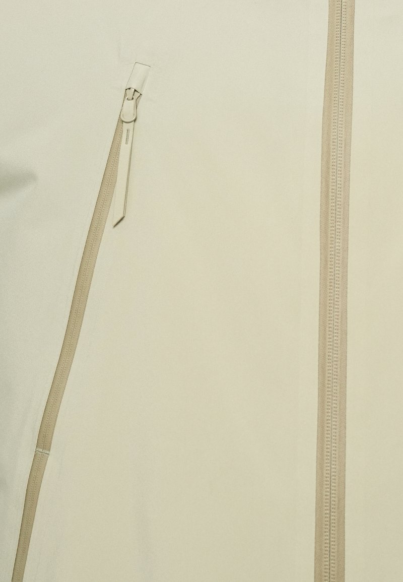 Veste beige clair avec une texture lisse, une fermeture éclair centrale bien visible et des détails de zipper beige contrastants sur le devant.