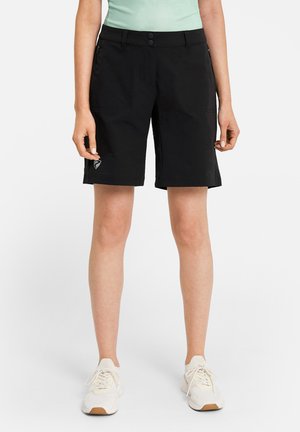 Person trägt schwarze knielange Shorts, hellgrünes Oberteil und weiße Turnschuhe, steht vor einem schlichten weißen Hintergrund.