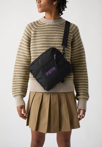 Juodas Jansport per petį nešiojamas krepšys su užtrauktuku, stačiakampio formos ir reguliuojama petneša, dėvimas ant dryžuoto megztinio ir klostuotos sijono.