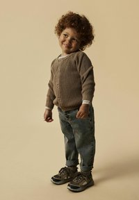 Brauner gestrickter Cardigan mit Knöpfen, weißes Unterhemd, hochgekrempelte blaue Jeans und schwarze/braune Sneaker mit strukturierter Oberfläche.