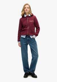 Sudadera burdeos con cremallera, capucha con cordón, bolsillos frontales y detalle de logo, combinada con una camiseta de color azul claro y jeans azules holgados. Zapatos negros.