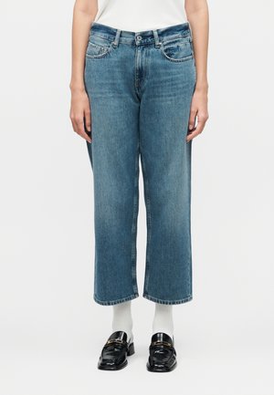 Person som har på seg blå cropped jeans med rette ben, hvite sokker, svarte loafers med gull-detaljer og en hvit ermeløs topp.
