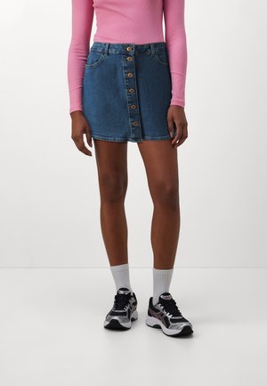 Falda de mezclilla con un diseño ajustado, color azul, cierre frontal de cinco botones y dos bolsillos laterales. Combinada con una blusa de manga larga rosa y zapatillas deportivas.