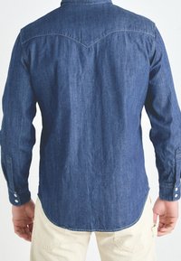 Long-sleeve denim shirt in dark blue, featuring a curved yoke, buttoned cuffs, and a slightly rounded hem for a relaxed fit.