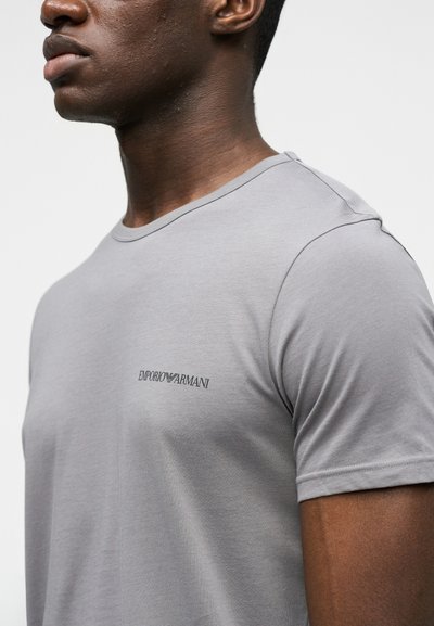 Κοντινό πλάνο ενός άνδρα που φορά ένα ανοιχτό γκρι T-shirt με μικρό λογότυπο "Emporio Armani" στο αριστερό στήθος, σε απλό φόντο.
