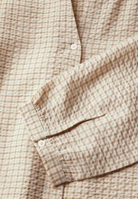 Camicia a quadri beige chiaro realizzata in un tessuto testurizzato con piccoli motivi a scacchi, dotata di bottoni bianchi e dettaglio con maniche ripiegate.