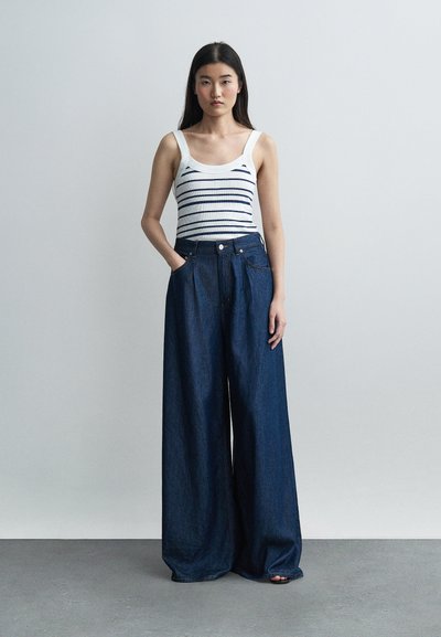 Young woman standing wearing a white sleeveless striped top and high-waisted wide-leg dark denim pants against a plain light background.