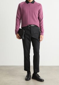 Homme portant un pull violet côtelé avec col noir, pantalon noir, ceinture noire, chaussures en cuir noires, tenant une pochette noire.