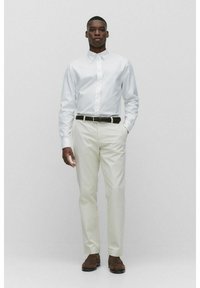 Chemise blanche à boutons avec des manches longues, pantalon crème, ceinture tressée marron et chaussures marron. Le mannequin se tient avec les mains dans les poches.