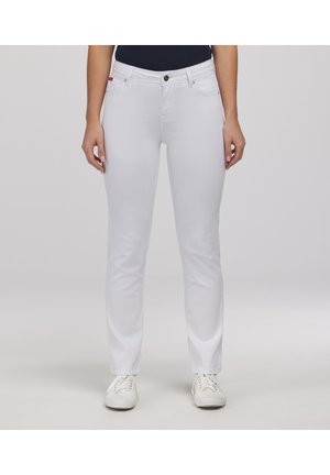 Kobieta w dopasowanych białych dżinsach z przednimi kieszeniami i białych trampkach, stojąca na tle jasnego, jednolitego tła.