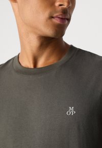 Close-up of a person wearing a dark gray crew neck t-shirt with "M O'P" white logo on the left chest.