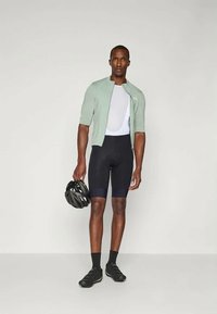 Cyklistický odev pozostávajúci z svetlozeleného trička s krátkym rukávom, bielej sieťovanej spodnej vrstvy, čiernych šortiek a čiernych cyklistických topánok, s prilbou v ruke.