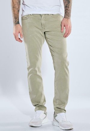 Man wearing beige slim-fit jeans, white sneakers, and a white t-shirt, standing against a plain light background.