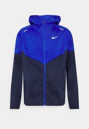 Chaqueta cortavientos azul y azul marino con capucha de Nike, con cremallera frontal y logo blanco en el pecho, diseñada para uso ligero en exteriores.