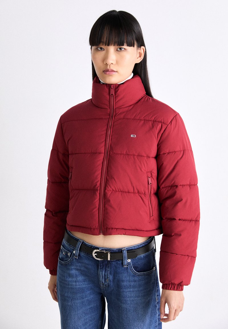 Röd kort puffjacka med hög krage, med stygnsektionsdetaljer, dragkedja framtill och en ficka på vänster sida. Bärs över blå jeans.